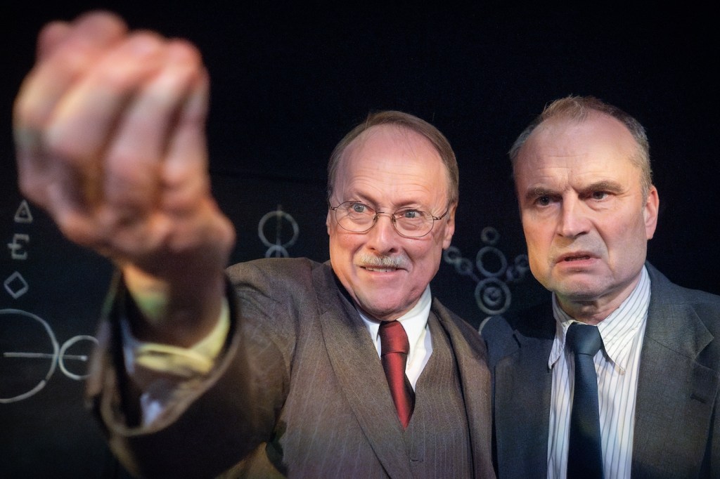 Stephen Riddle as Carl Jung standing next to Jeremy Drakes as Wolfgang Pauli and extending his arm