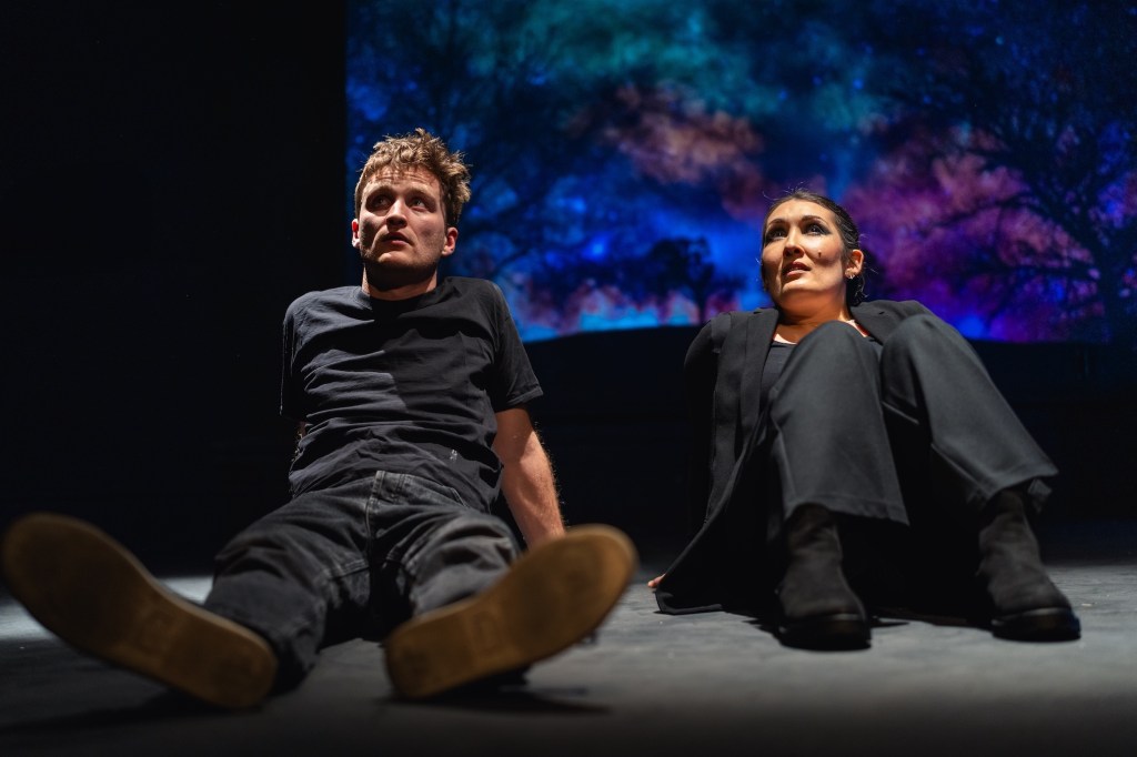 Conor Rowlett as Charlie and Emily O'Mahony as Tango dressed in black, sitting on the ground looking up, with a dark landscape behind.