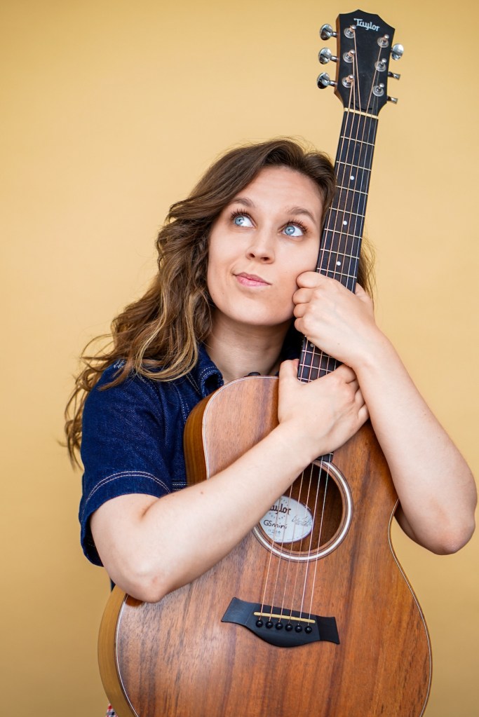 Evelyn Rose holding an upright acoustic guitar and looking thoughtfully up to her right
