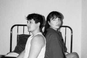 Black and white photo. David (Boyan Petrov) and Phoebe (Lyndsey Ruiz) back to back on a bed. He in white sleeveless vest, she in dark top. He is looking to the left, she is looking straight ahead towrads the camera.
