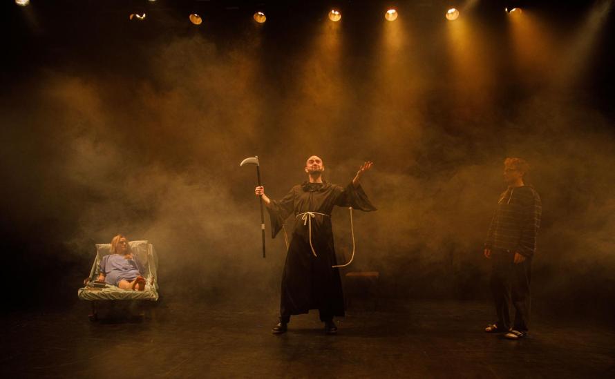 On the right Joe Bellis as Death, arms raised; on the left, Hannah Harquart, lying in bed in a blue hospital gown.