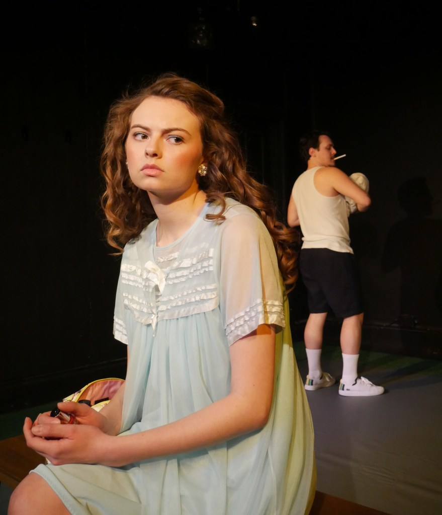 Marianne (played by Halli Patterson) in the foreground pensively seated on the left wearing a pale blue filly nightdress; Peter (played by Boyan Petrov) in the background on the right dressed in vest and shorts, holding their bay and smoking