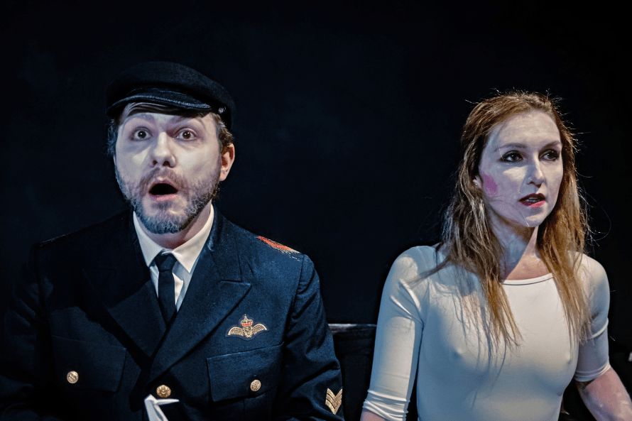 Seated on the left Daniel Chrisostomou in uniform cap, shirt, tie and militaristic jacket; seated on the right Vyte Garriga, white makeup and white t-shirt.