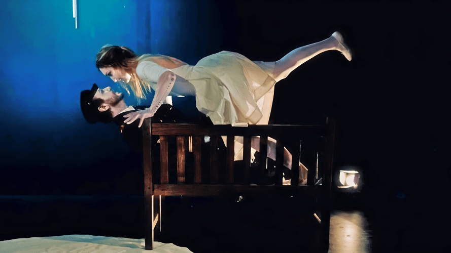 Vyte Garriga in a white ballerina costume leaning menacingly over reclining uniformed Daniel Chrisostomou on a park bench