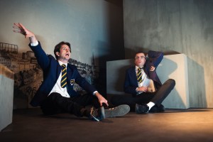 On the left Paddy (played by Eoin Sweeney), seated cross-legged in school uniform of white shirt, black trousers, purple blazer with crest and purple and yellow striped tie. He is gesticulating wildly with his right arm raised. On the right is Mick (played by Matthew Blaney) also cross-legged and wearing the same school uniform. He is wearing glasses and looking terrified.