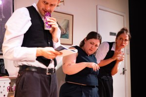 On the left Malcolm (played by Gabriel Fogarty-Graveson); in the middle Harriet (played by Laurel Marks); on the right Regina (played by Gabrielle Nellis-Pain). They are all wearing dark undertaker-style clothes and looking quizzically at something off camera to their right.