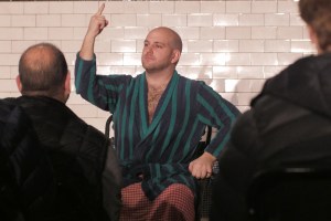 Nick Cassenbaum, seated, wearing a green and blue striped dressing gown, one hand pointing upwards