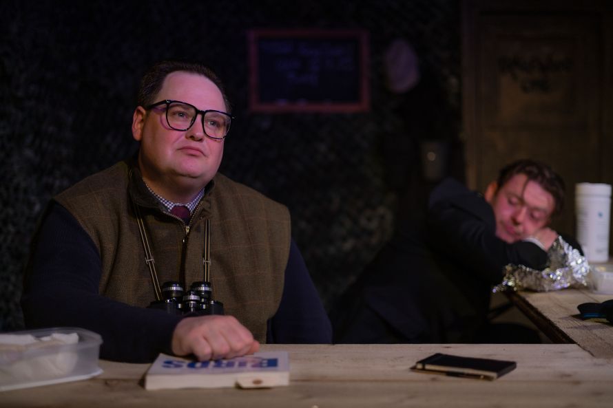 On the left Roy (played by Jack Robertson) gazing out in bird-watching gear and binoculars; on the right Dave (played by Calum Lill) out of focus, eyes closed, slumped down head on the table.