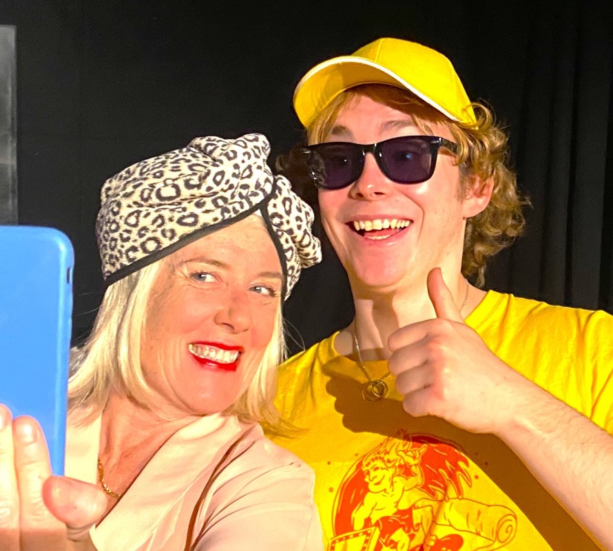 On the left Ruth Madoff (played by Emily Swain), wearing a leopard-print headscarf and grinning, taking a selfie with pizza delivery boy Marco di Cesare (played by Evan Emanuel) wearing a bright yellow and red t-shirt, yellow hat and dark sunglasses, giving a thumbs up.