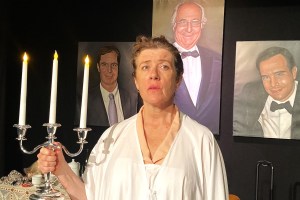 Ruth Madoff (played by Emily Swain) dressed in a white dress and holding a lit three-pronged candelabra. Behind her are paintings of her husband and two sons.