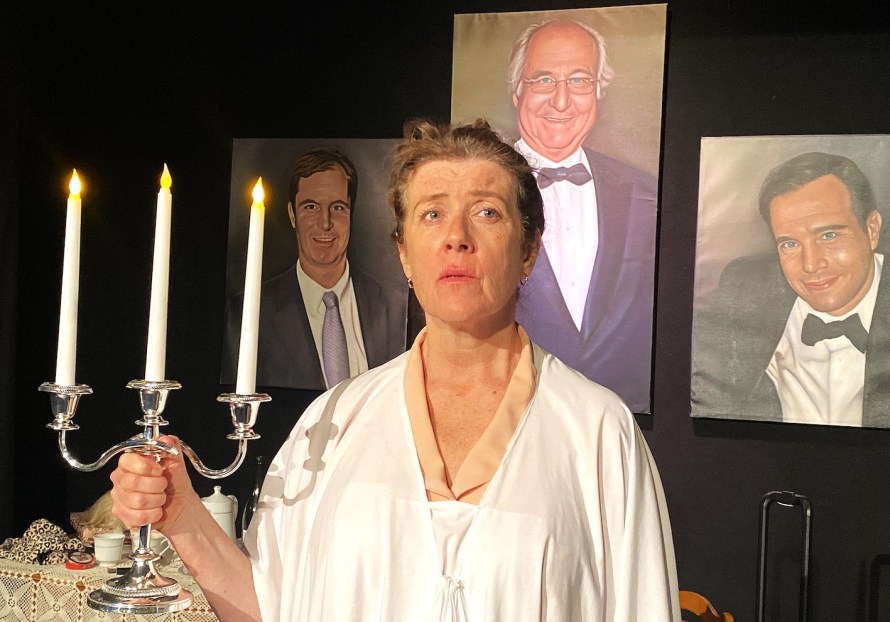 Ruth Madoff (played by Emily Swain) dressed in a white dress and holding a lit three-pronged candelabra. Behind her are paintings of her husband and two sons.