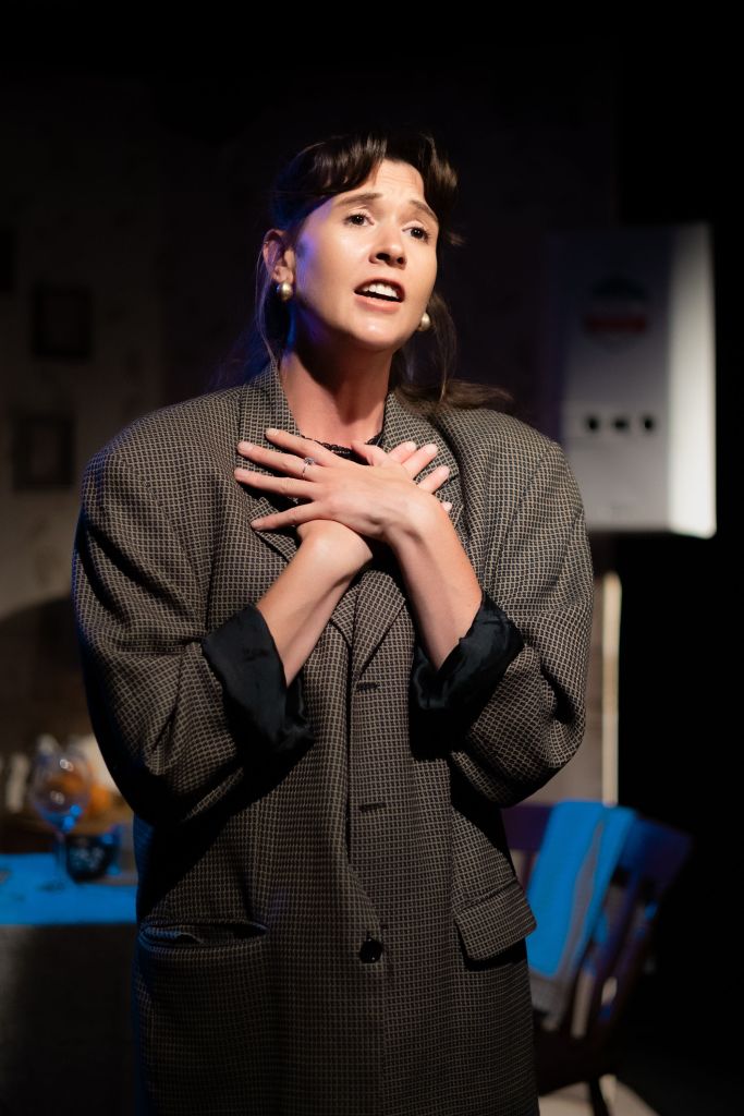 Young Nadine (played by Ellie Baldwin), wearing a check jacket, standing and singing, with hands spread over her chest.