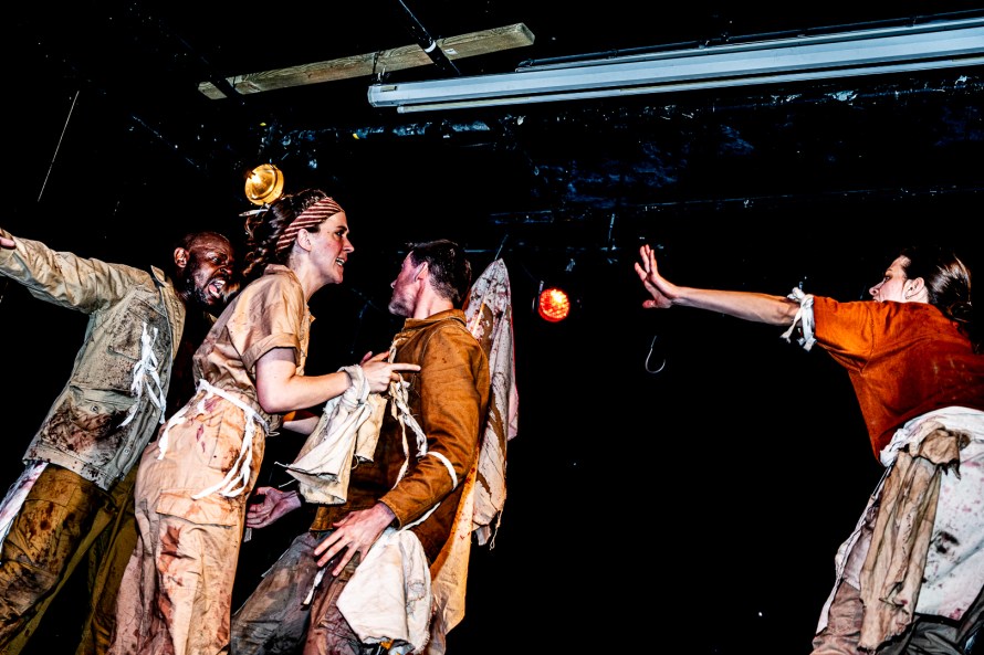 Left to right: Arnold Patrick Lumu standing shouting and gesticulating; Greta Hansen standing and pointing to her right; Louis Cruzat turned away to his right; distanced Clio Carrara arm outstretched towards the other three.