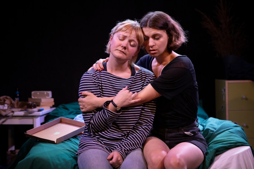 On the left, sitting with eyes closed on a messy bed, Katie (played by Catherine Ashdown), wearing a horizontal stripy blue and white top and jeans; sitting on the right, with arms round Katie, Roni with eyes closed (played by Eileen Duffy) in black shorts and T-shirt; on the right,