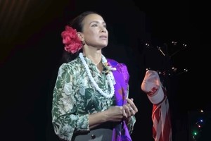 Ann (played by Nikol Kollars) wearing an open-necked green flowery blouse, with a red flower in her hair, a white necklace and purple sash, standing and about to sing.