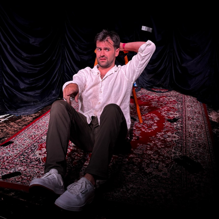 Blake Stratso seated on the floor, resting against a bar stool in open-necked white shirt, tan chinos and white trainers. There is an Indian rug on the floor. He is looking to the side and pointing forwards.