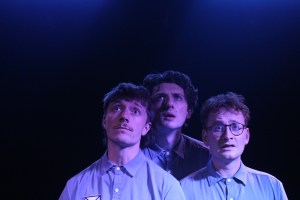 Bathed in a blue light, on the left Gavin (played by Sam Plumbe), in the centre Clint (played by Ewan Bruce) and on the right Brian (played by Henry Calcutt). They are all wearing corporate uniforms and gazing nervously towards an unseen screen.