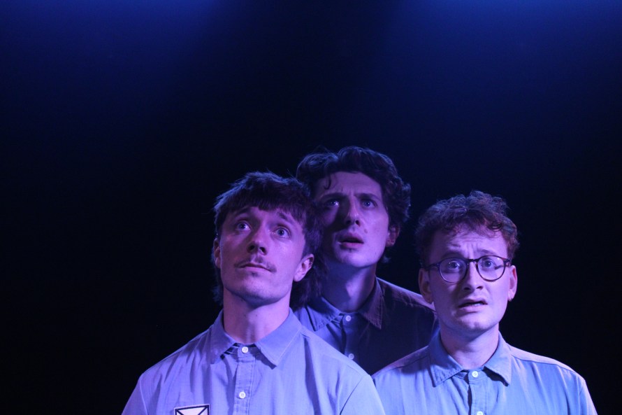 Bathed in a blue light, on the left Gavin (played by Sam Plumbe), in the centre Clint (played by Ewan Bruce) and on the right Brian (played by Henry Calcutt). They are all wearing corporate uniforms and gazing nervously towards an unseen screen.
