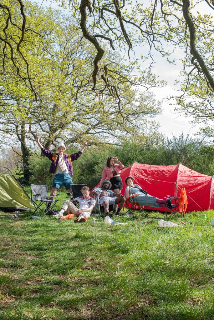 A woodland setting with a green tent on the left and a red tent on the right. Five characters ranged in front of the tent in Festival mood. Left to right are Marvin (played by Alistair Rowley), Jordan (played by Tom Feasby), Jason (played by Tafadzwa Phillip Madubeko), Daisy (played by Georgia Dye), and Lisa (played by Marnie Yule).