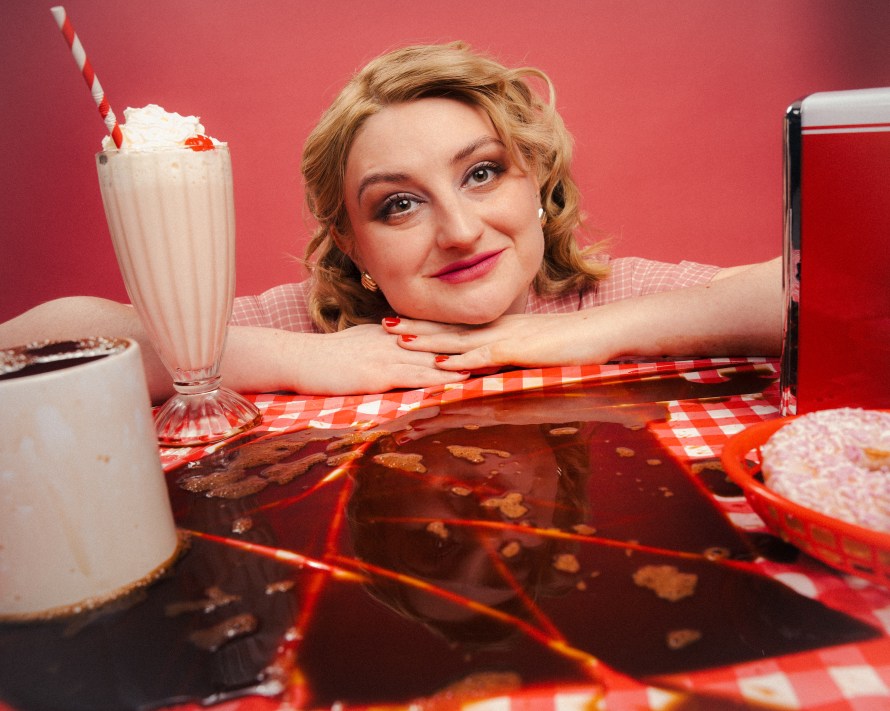Lorna Rose Treen, head resting on her hands, looking over a diner-style table with milkshake and coffe cup.