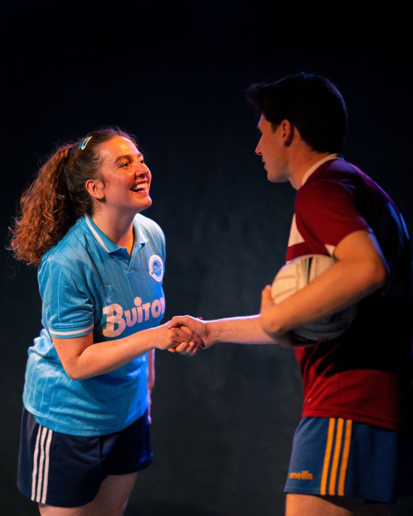 On the left, in light blue football kit, Melissa (played by Dión Di Maio) shaking hands with Deren (played by Jake Douglas) on the right also in football kit and holding a Gaelic football.