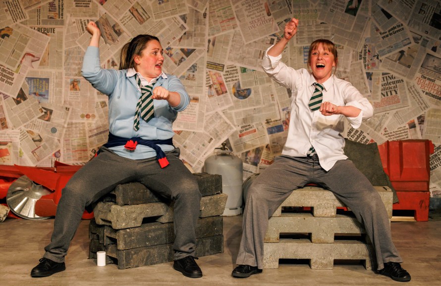 Two teenage girls in scruffy school uniforms sitting on concrete blocks and riding imaginary horses.On the left Zoe (played by Lauren Downie) and on the right Kay (played by Abigail Hood)