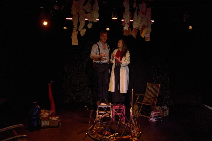 Standing on a stoll on the right El (played by Heather Jones) in a long cream mac over a red top and dark trousers; standing on a higher stool on the left, wearing a pale blue shirt and dark trousers with braces is Tophe (played by Stanley Eldridge). They are facing a small fire surrounded by a frame of branches.