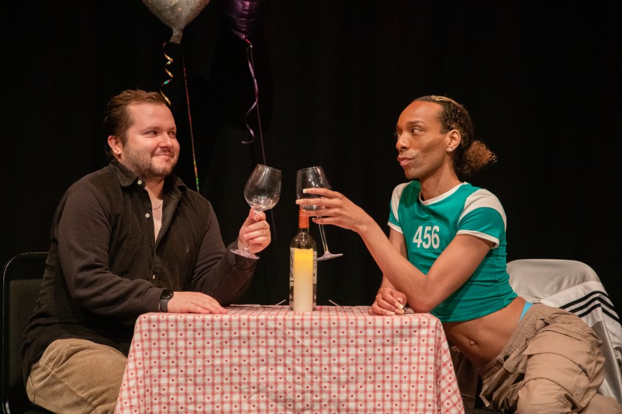Two men seated at a restaurant table. On the left, Ben, played by Sam Danson, in a dark shirt and beige cords; on the right Tim, played by Dior Clarke, in a midriff-revealing turquoise T-shirt and cargo pants. They are toasting each other with wine glasses.