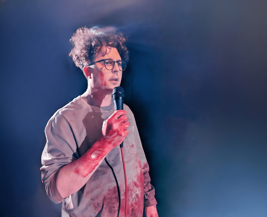 Olly Hawes, standing up holding a microphone, bloodied arms, blood-stained grey sweatshirt