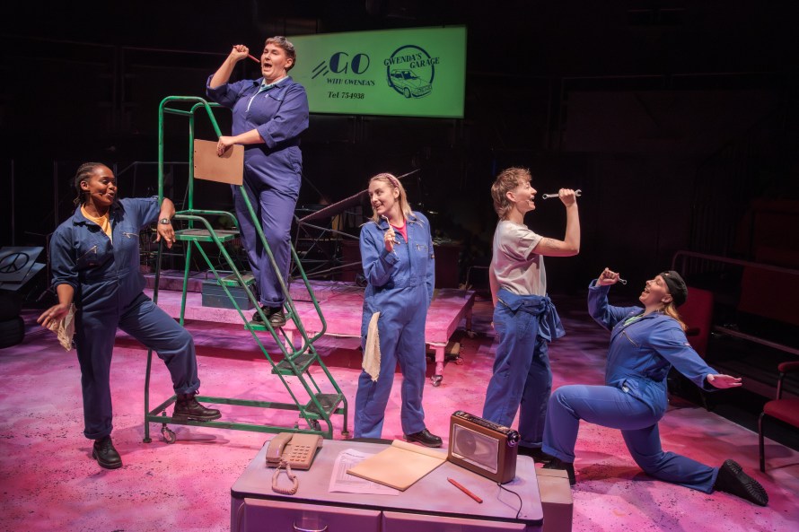 A group of female mechanics dressed in blue overalls arranged in a dance group. Left to right - Terry (played by Sia Kiwa), Carol (played by Eva Scott), Bev (played by Nancy Brabin-Platt), Dipstick (played by Lucy Mackay) and Dance Captain Georgina Coram
