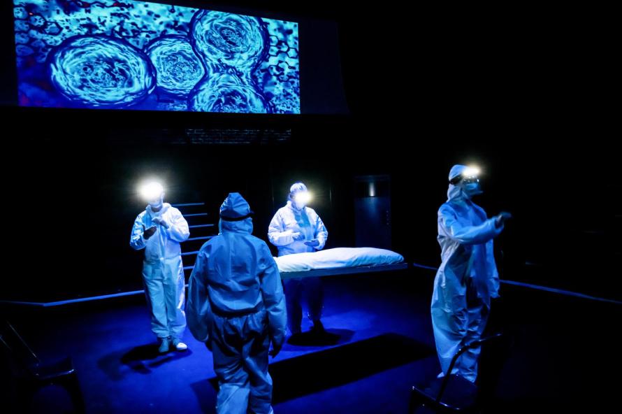 In blue light, four surgeons in white hazmat suits and with head torchs shining surround a suspended surgery bed. A projection of blue dividing cells hangs above the bed.