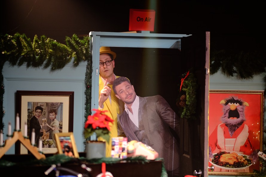 In a Christmas home themed TV set - behind the door is Tom Clarkson in bright yellow suit, yellow tie and yellow bowler hat. In front of him is life-sized cutout of Duncan from Blue.