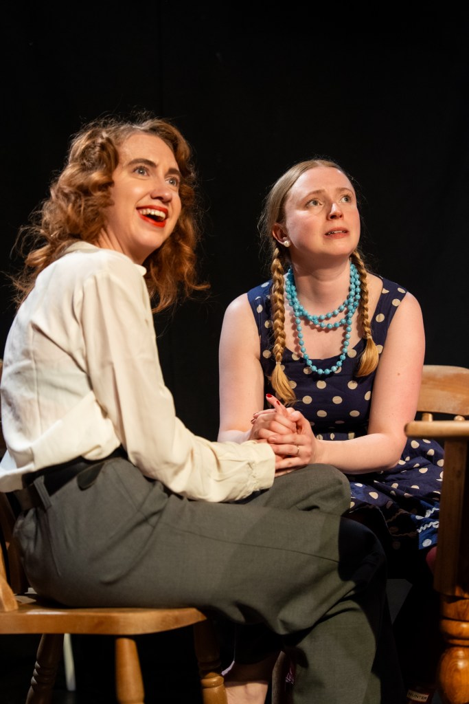 Seated on the left Gloria Wilder (played by Dany Van Brabant) in white blouse and grey trousers, smiling; seated on the right is Martha Hueber (played by Evie Weldon) in blue dress with white polka dots and a pale blue beaded necklace.
