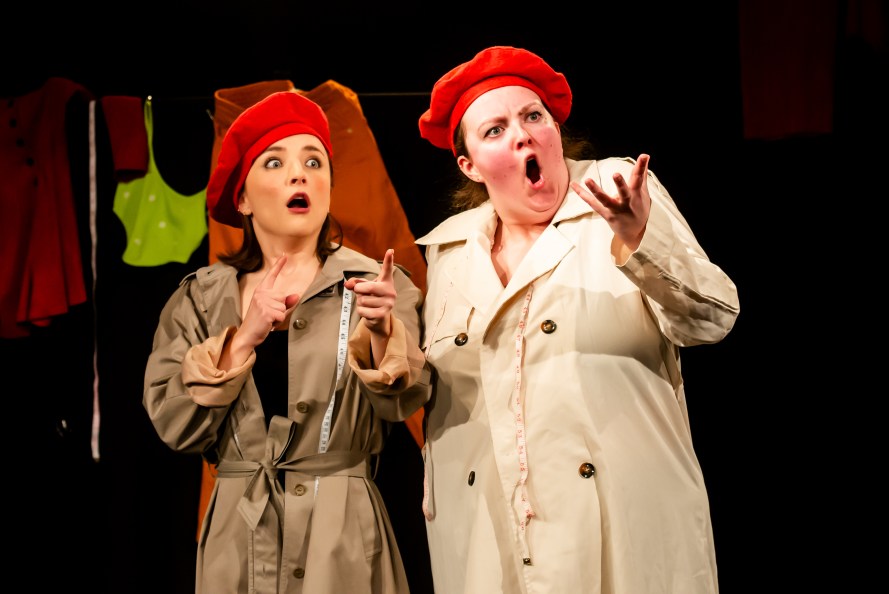 Standing on the left Oche (played by Kiera Murray) wearing tan raincoat, red beret and tailor's measuring tape; standing on the right, in matching outfit is Bree (played by Hannah Mcleod). They are gesticulating and looking outraged.