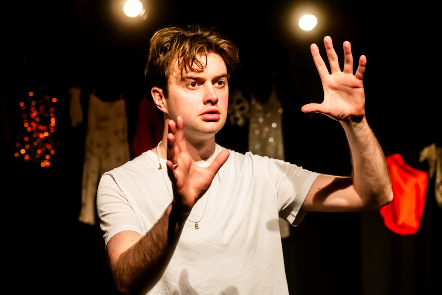 Ze Emperor (played by Jacob Baird) in white T-shirt and hands raised, looking forwards.