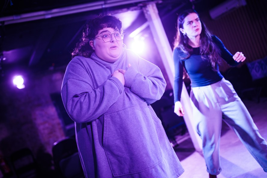 Standing on the left Bridget (played by Beca Barton) in a grey hoodie; standing on the right in black top and grey trousers Sally (played by Mimi Millmore); both angrily addressing someone off camera to the right.