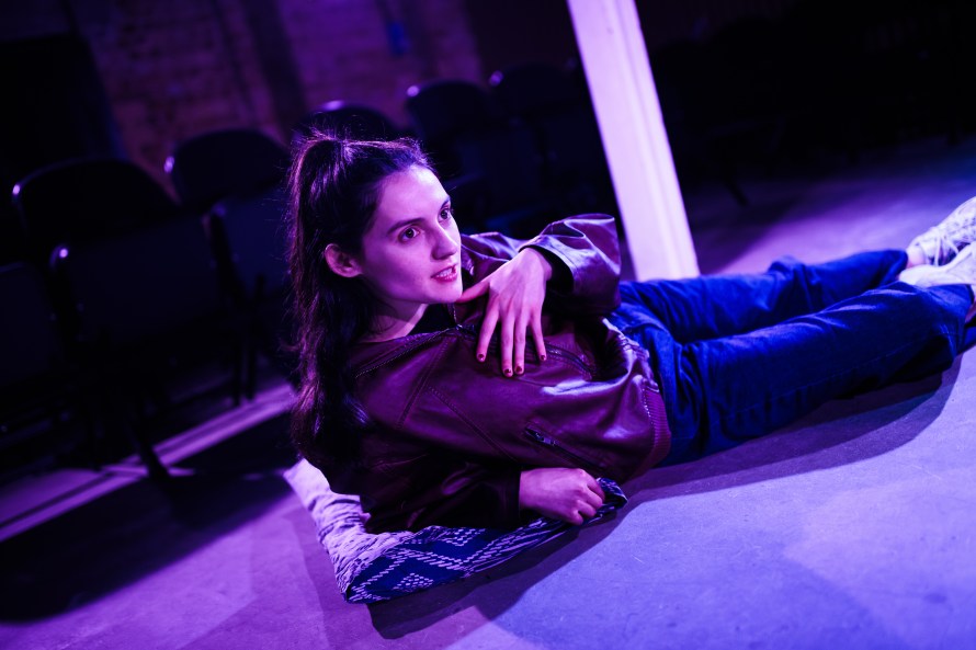 Woman (played by Anna Coupe) in dark leather jacket, blue jeans and trainers, lying on the floor and leaning on a cushion, with her hand argumentatively on her chest.