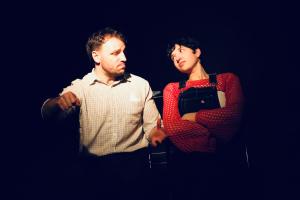 On the left, in open-necked check shirt Gary (played by Charlie Jennings), on the right, looking at sceptically, dressed in red crocheted top and blue dungarees, Sam (played by Gaby Coleman).