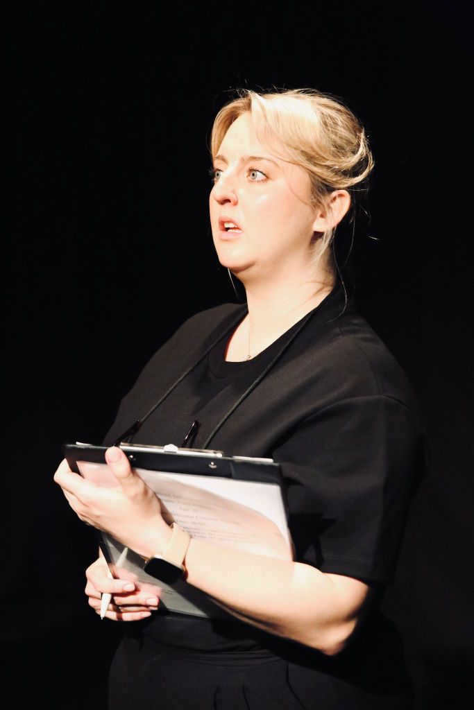 Standing in black, holding a clipborad and pen, blond hair severely combed back, the Chaperone (played by Lucy Forker)
