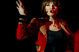 Sarah McGuinness in red and gold ringmaster style jacket and black vest, gesticulating.