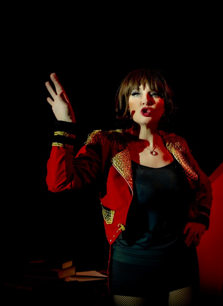 Sarah McGuinness in red and gold ringmaster style jacket and black vest, gesticulating.