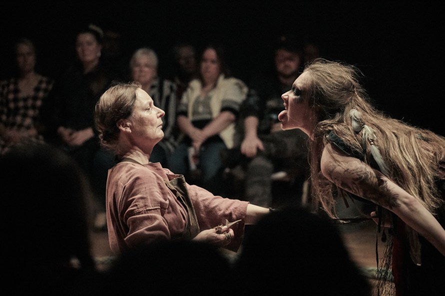 Muted brown colourscape, on the left Widow Pruttock (played by Emily Outred) holding a small knife towards Sheldis (played by Bess Roche) who is lunging towards her with tongue sticking out.
