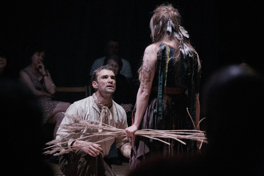 Muted brown colourscape, seated on the left Man Billy (played by Mylo McDonald); standing on the right with her back to us holding a bunch of reeds and looking intently towards him is Sheldis (played by Bess Roche).