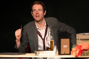 Craig Kitman (played by Benjamin May) seated at a messy desk in grey jacket, white shirt and loosened red tie, holding a fountain pen and looking messianic.