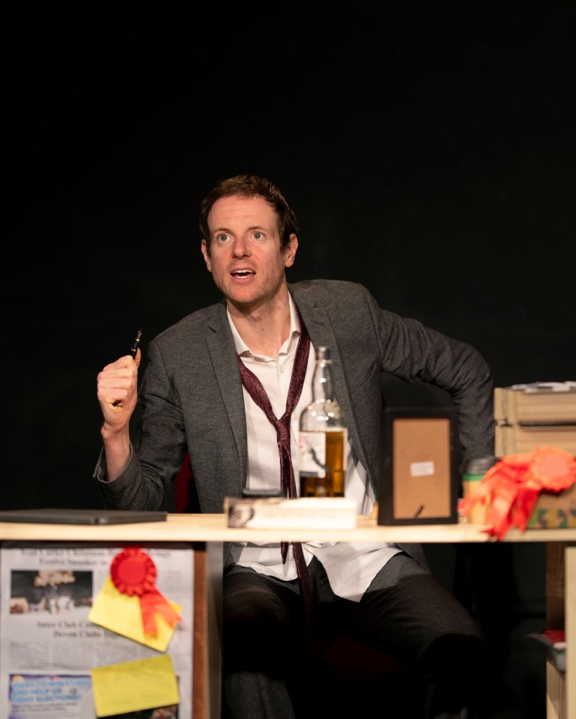 Craig Kitman (played by Benjamin May) seated at a messy desk in grey jacket, white shirt and loosened red tie, holding a fountain pen and looking messianic.