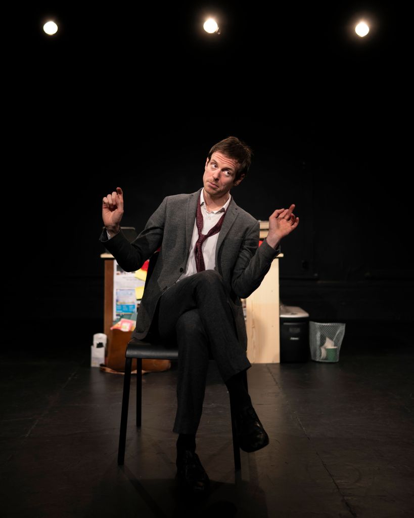Craig Kitman (played by Benjamin May) seated cross legged on a chair in grey suit, white shirt and loosened red tie, holding out his hands in a silly Mr Bean-like pose.